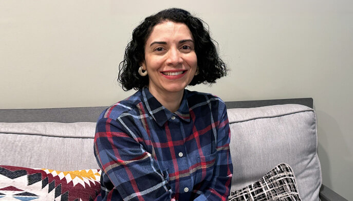 Claudia Atehortua with short, curly black hair smiles while sitting on a gray couch. She wears a plaid shirt, with patterned cushions beside her, conveying a warm, relaxed atmosphere.